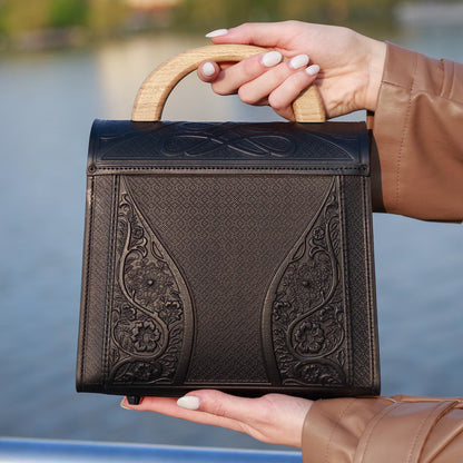 Person holding a brown leather handbag with intricate designs by a body of water.
