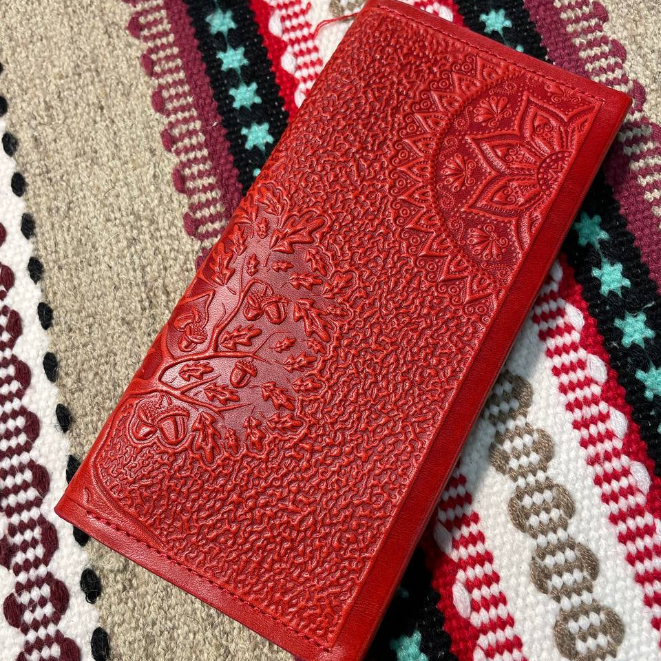 A red leather wallet with embossed floral pattern, placed on a fabric with a zigzag pattern. The background has a tribal-like pattern with stars.