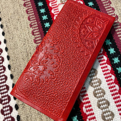 A red leather wallet with embossed floral pattern, placed on a fabric with a zigzag pattern. The background has a tribal-like pattern with stars.
